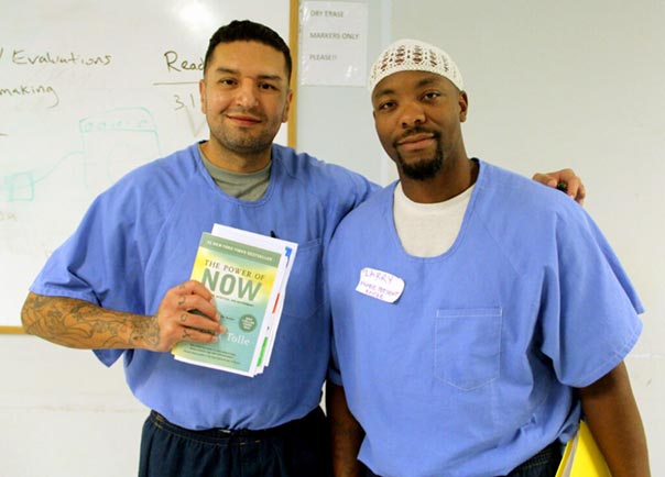 Two incarcerated men with arms around eachother smiling holding The Power of Now book by Eckhart Tolle